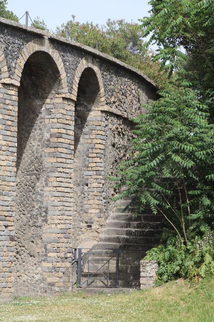 A stone brick viaduct with multiple arches extending across the image, set against a backdrop of greenery and a clear sky. To the right, a large leafy tree with vibrant green leaves partially obscures