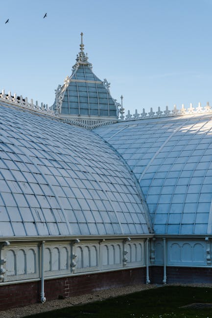 A detailed view of a historic glass and metal conservatory roof with intricate decorative trim and a prominent spire at the top, showcasing curved, segmented panes of glass supported by white-painted